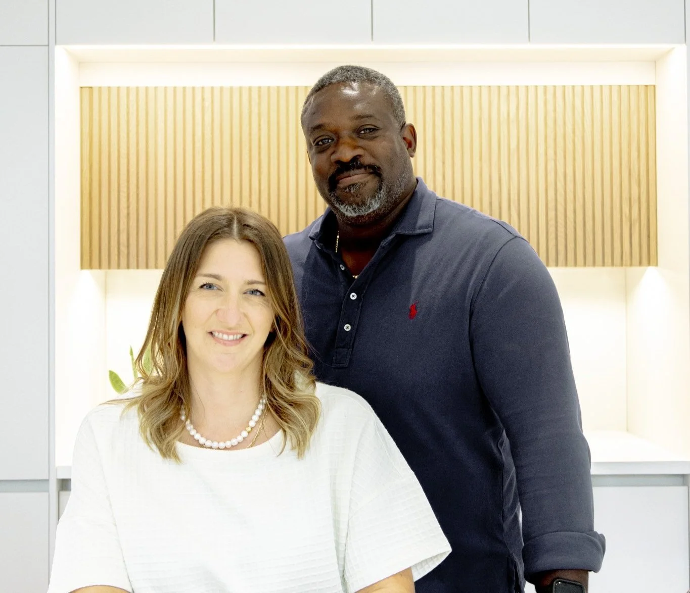 Julie has shoulder-length wavy hair wearing a white blouse and pearl necklace, sitting next to Chris with short gray hair and beard, wearing a navy blue polo shirt, in a bright modern room with wood paneling and white shelves.