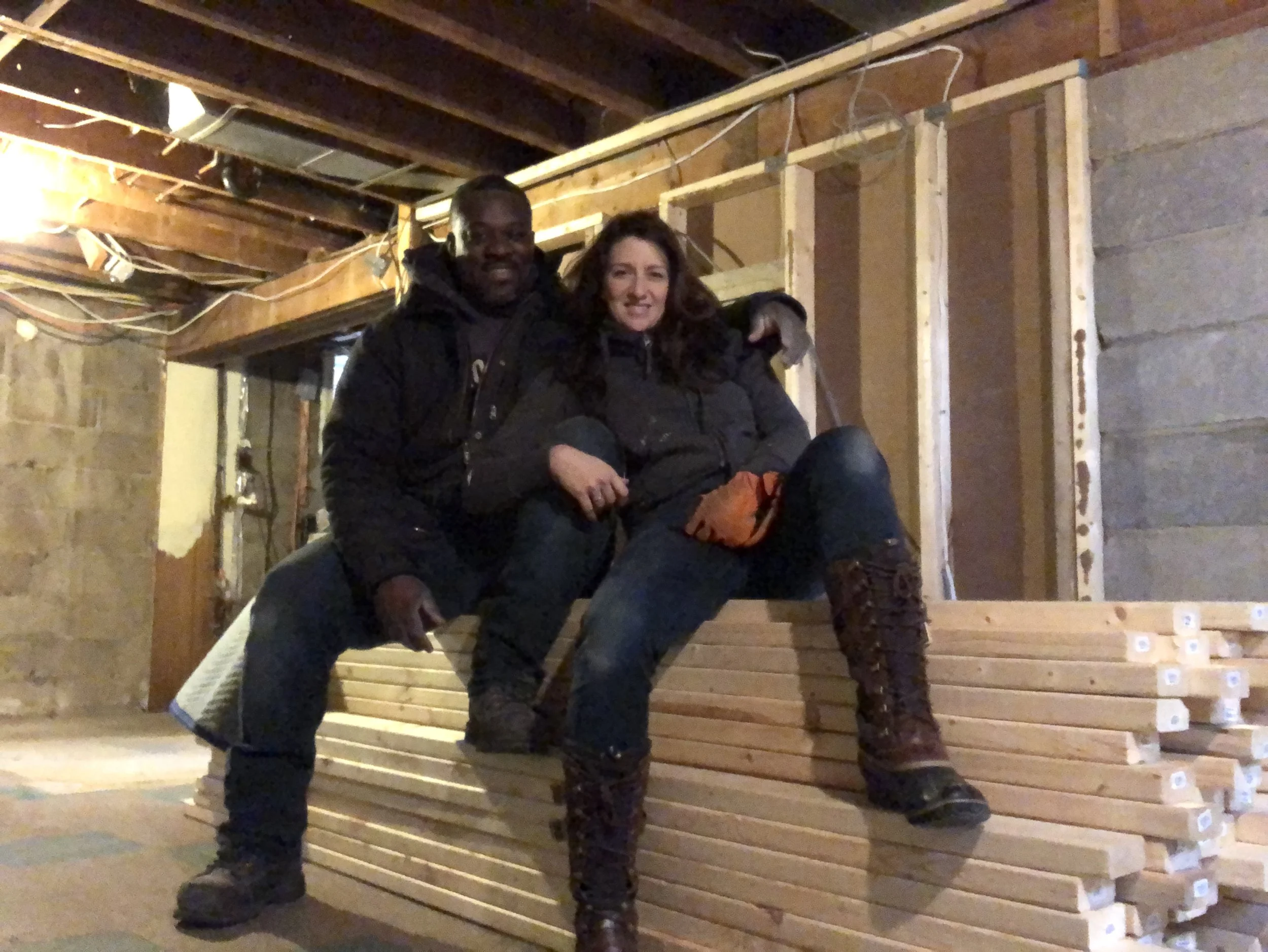 Julie and Chris, owners of Waterfront Home Improvement are sitting on a stack of lumber in a construction area, with unfinished walls and exposed wooden beams.