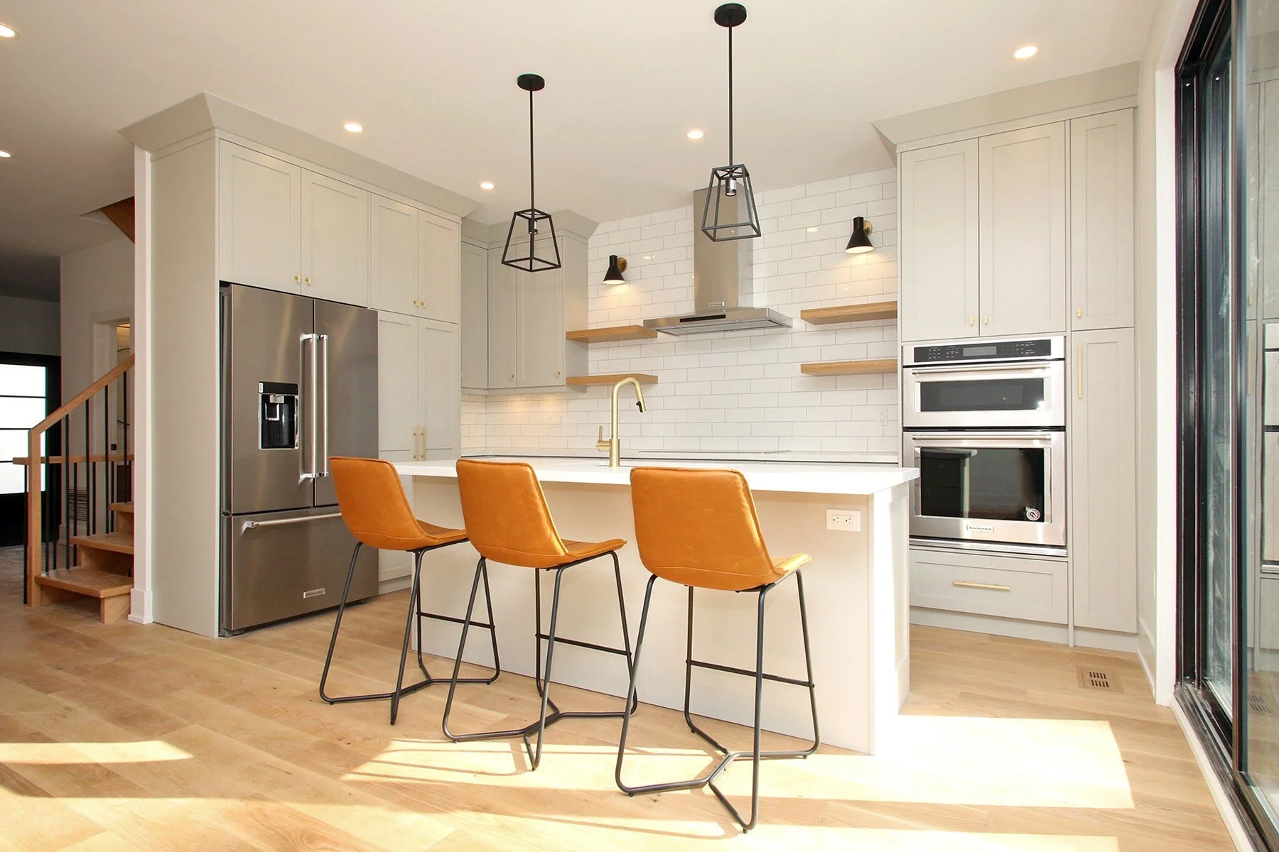 Modern kitchen with white cabinets, a large island, black pendant lights, orange bar stools, stainless steel refrigerator, double oven, and a sliding glass door.