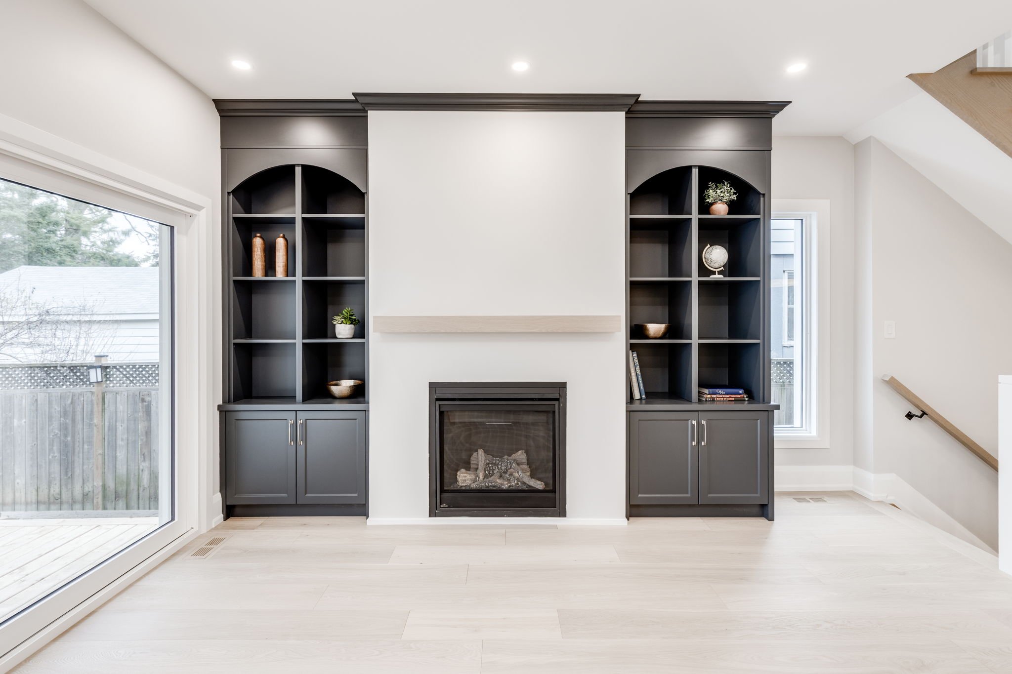 Living room after a whole-home renovation, with a gas fireplace flanked by charcoal arched built-ins, a white oak mantel, sliding doors to the deck, and the staircase beyond