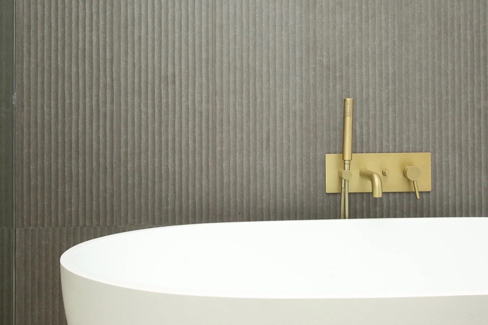 A modern white bathtub in front of a textured gray wall with a gold-colored wall-mounted faucet and handheld showerhead.