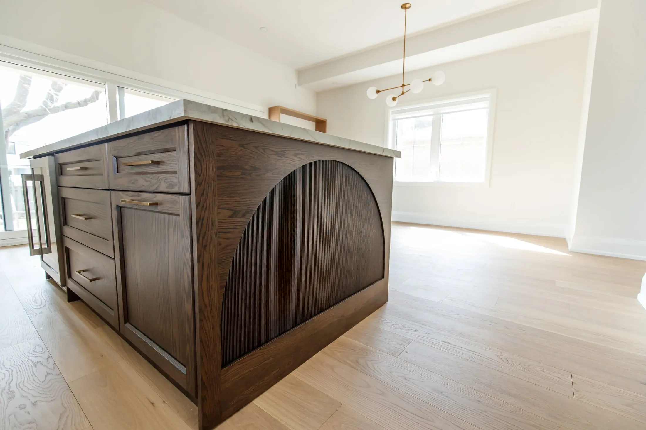 Kitchen island with an arched dark oak end panel and marble top, with a brass chandelier and wide-plank oak floors in the open dining area.