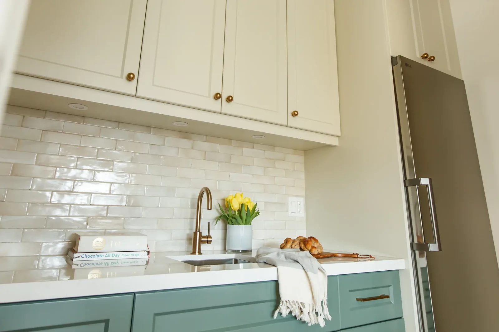 Teal base cabinets with cream uppers, a brass faucet, and a handmade zellige tile backsplash.