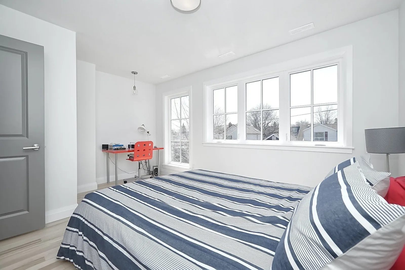 Bright white bedroom with large windows, grey door, light wood floor, and navy striped bedding.