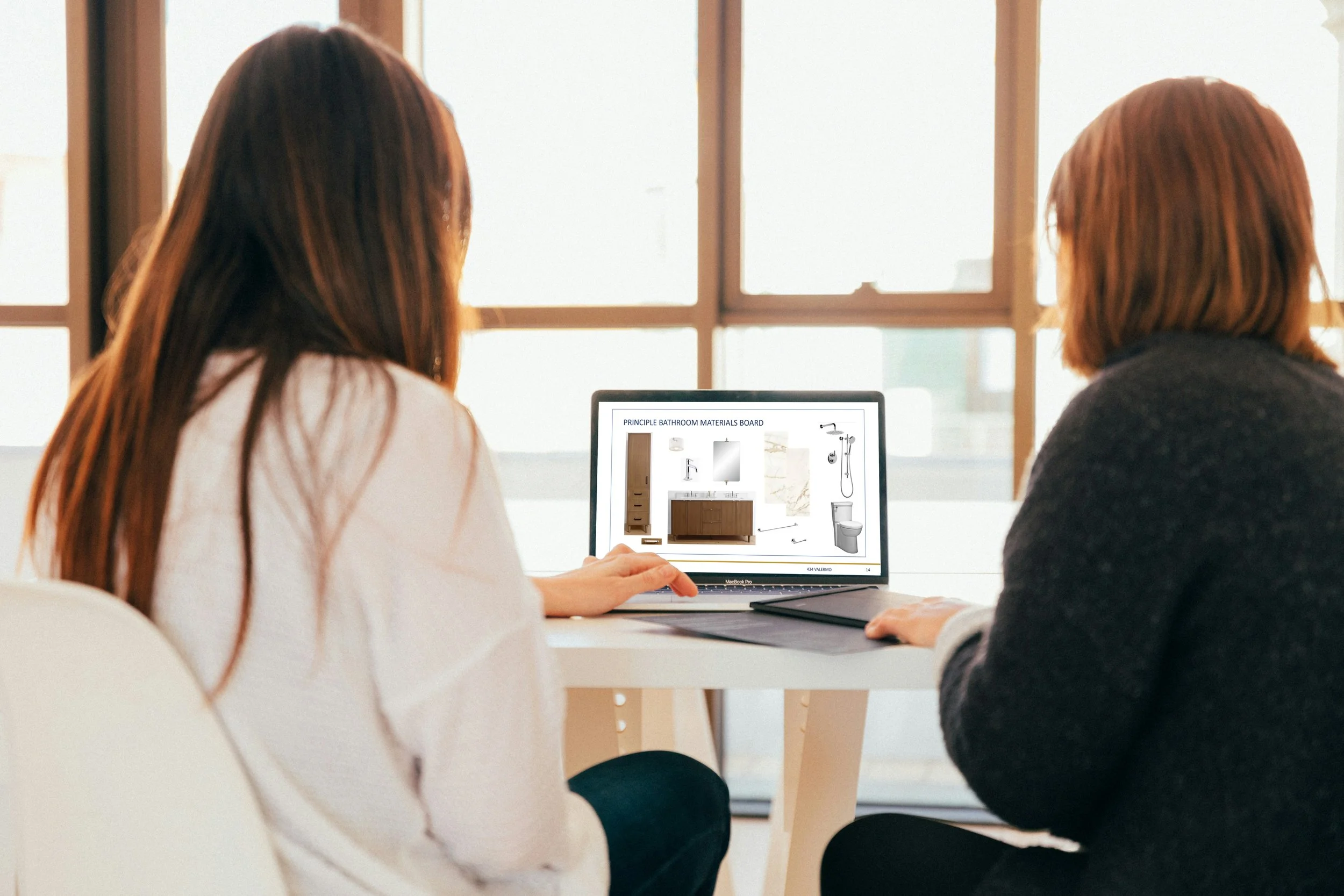 Two women sit at a table discussing bathroom materials displayed on a laptop screen, with a window in the background.