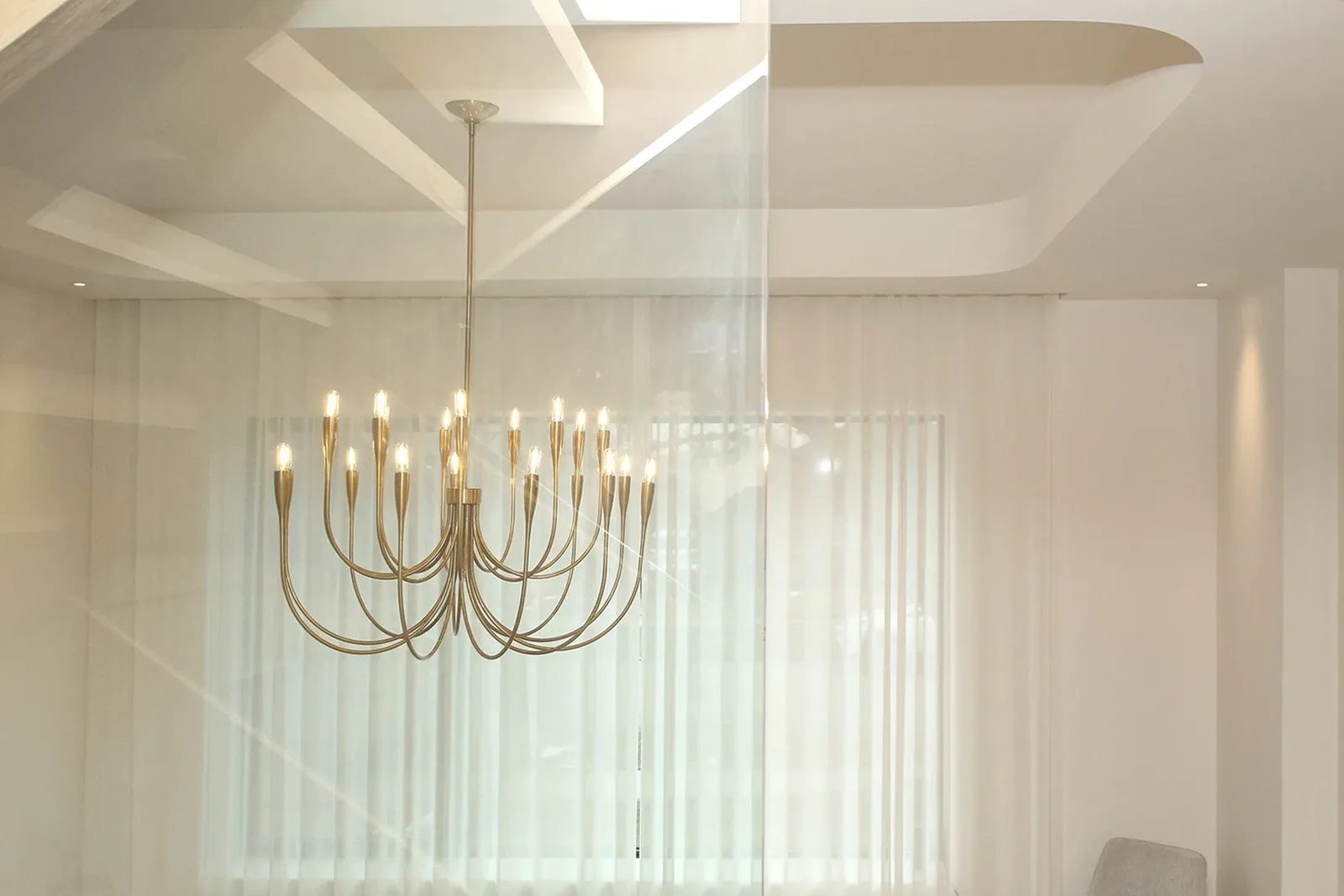 Close up of multi-arm satin brass chandelier beneath coffered ceiling with sheer linen drapery behind.