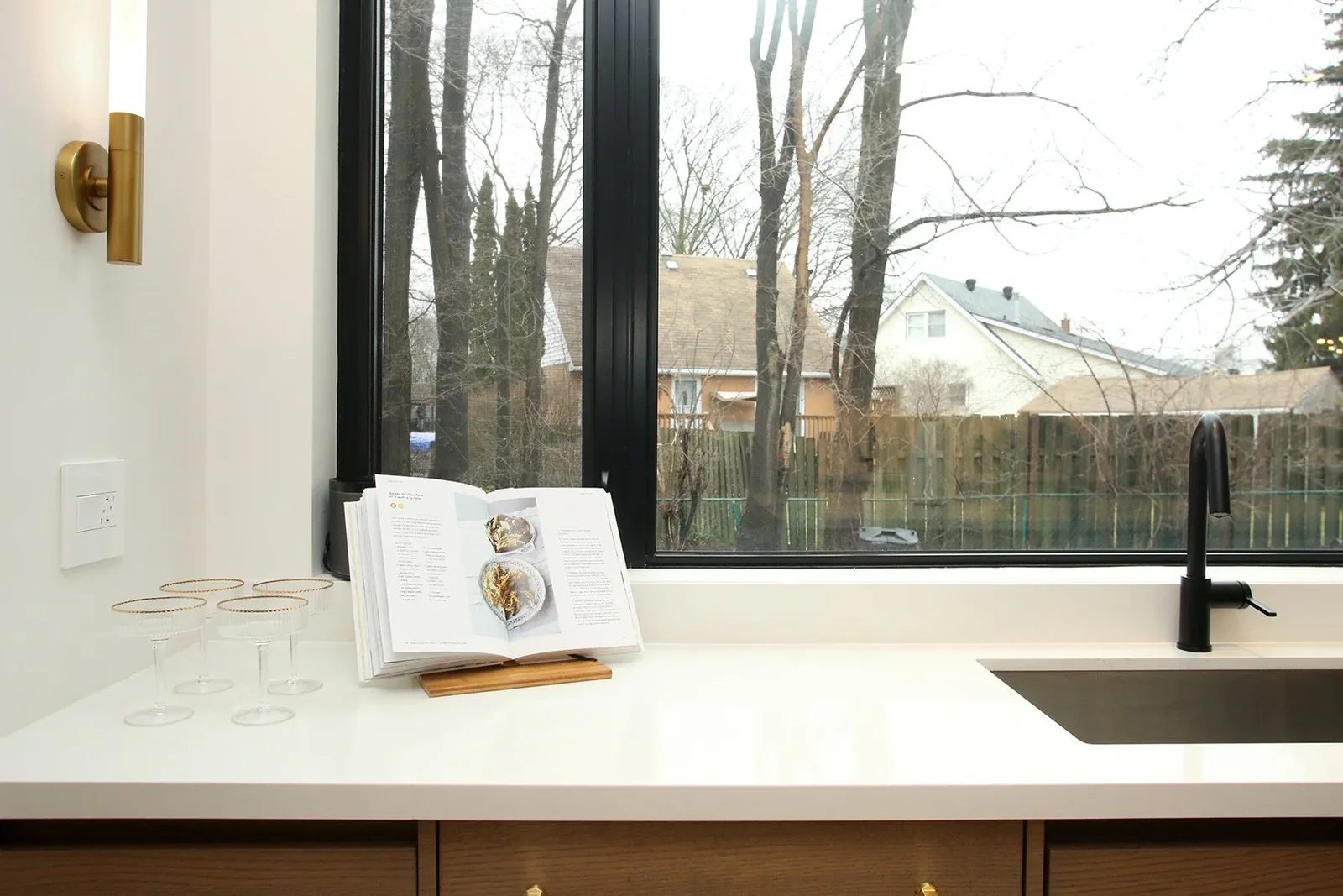 Undermount stainless sink and matte black pull-down faucet sit below a black-framed picture window with a brass sconce and coupe glasses styled on the counter.