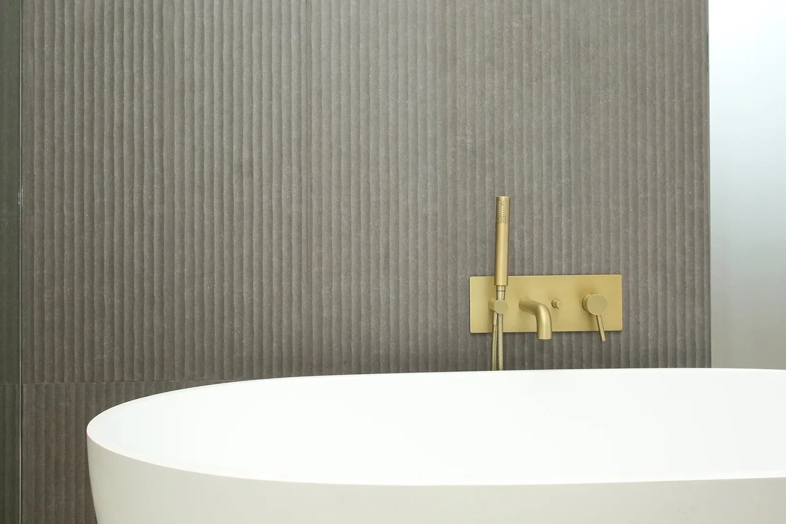 Detail of brushed brass wall mounted tub filler set against charcoal fluted tile feature wall.