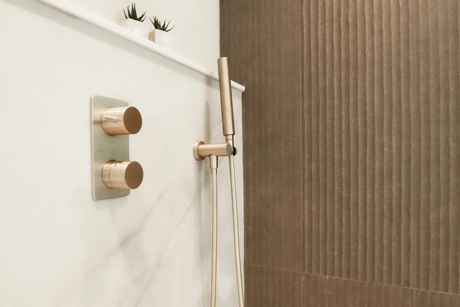 Close-up of a brushed brass thermostatic valve and handheld sprayer set against veined marble-look stone with a fluted taupe tile wall behind.