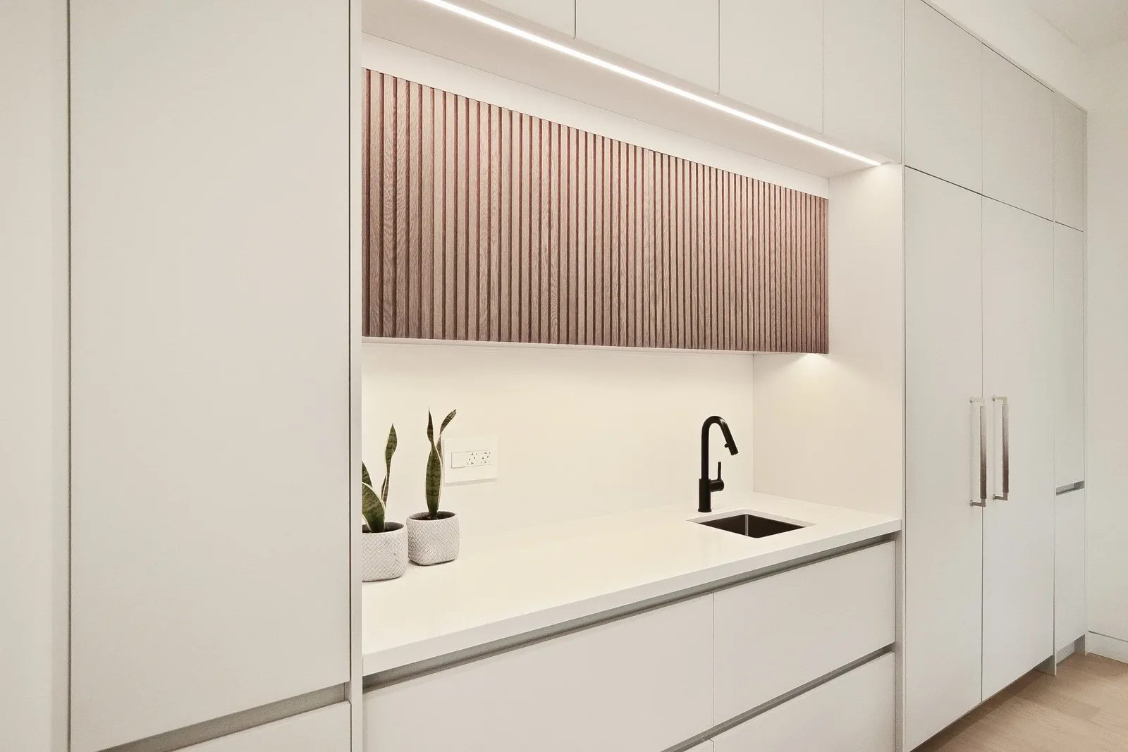 Minimalist servery nook with fluted walnut panel matte black faucet and handleless white cabinetry.