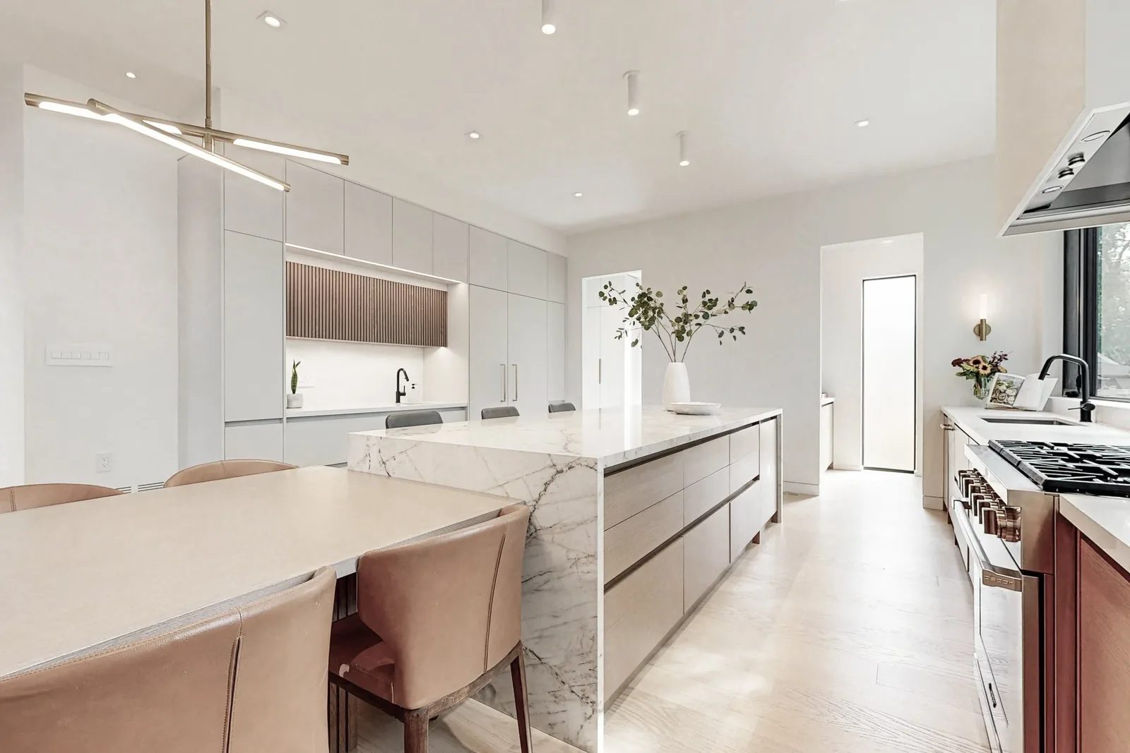Open concept kitchen and dining with oversized marble waterfall island light oak cabinetry and linear chandelier.