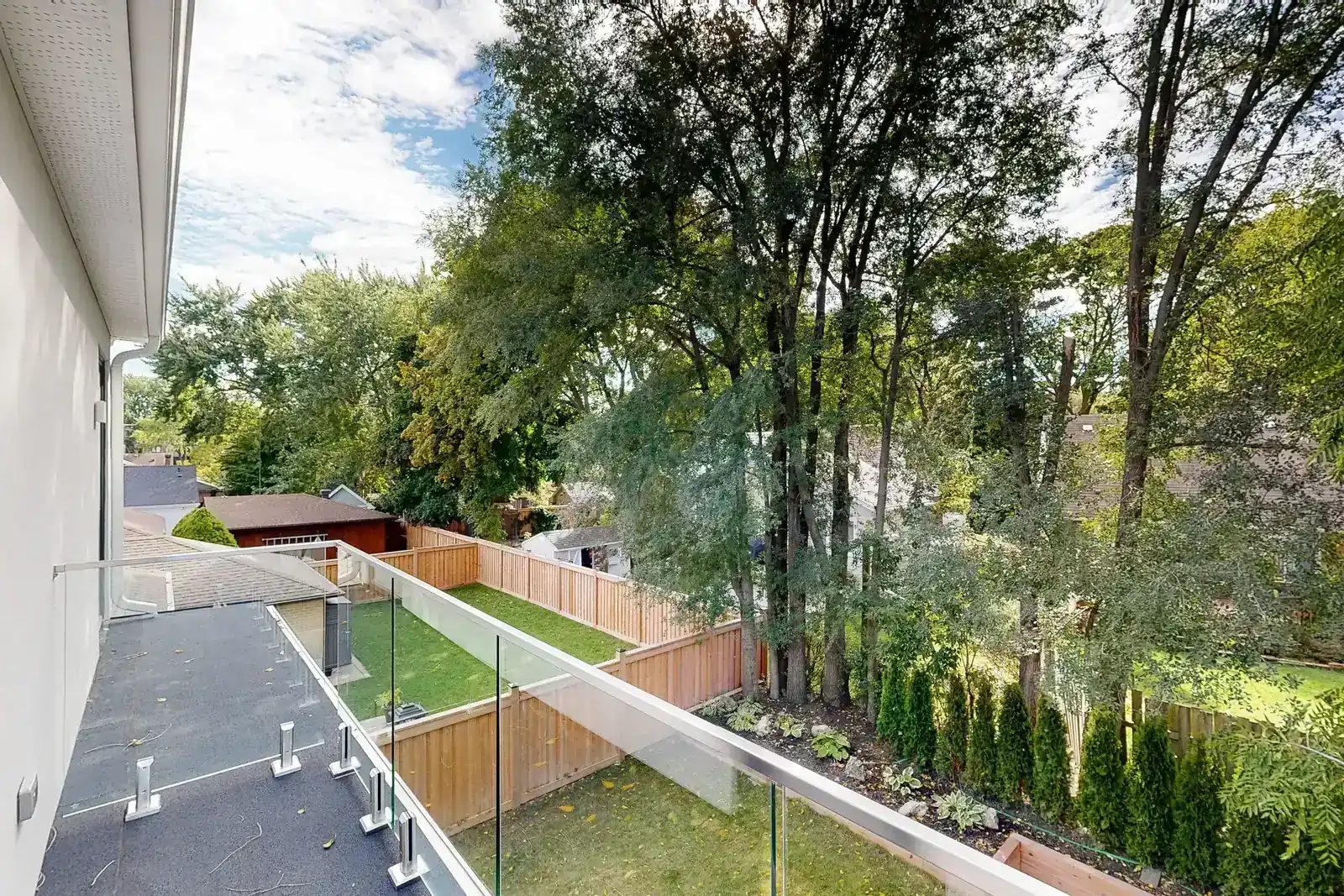 View from a second-storey balcony with frameless glass railing looking down over a landscaped backyard and cedar privacy fence.