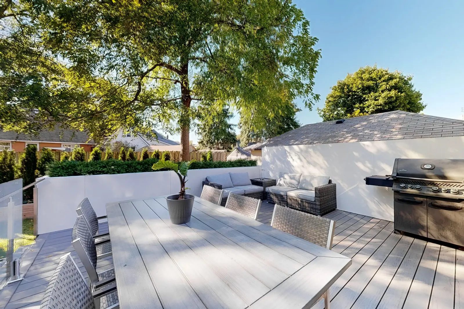 Rear deck with a stainless gas barbecue outdoor sectional and dining table framed by hedging and mature trees.