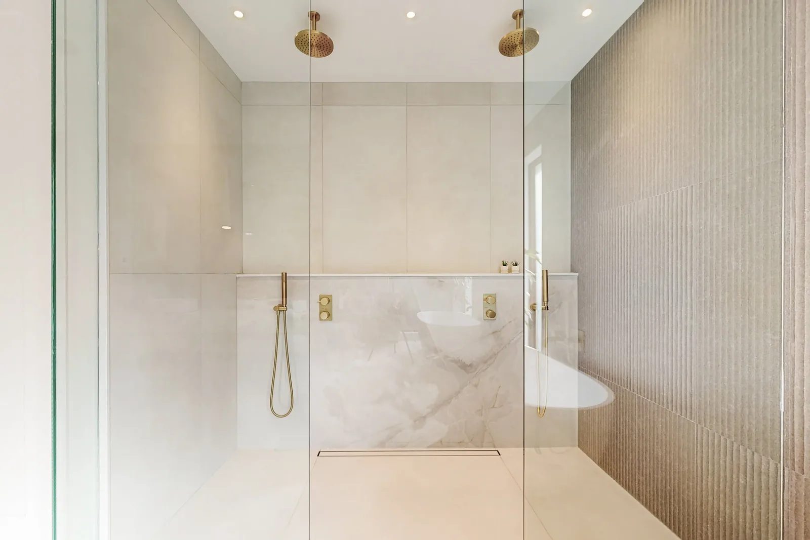 Double walk-in shower with two brushed brass rainheads handhelds and a fluted taupe tile accent wall opposite a polished stone wall.