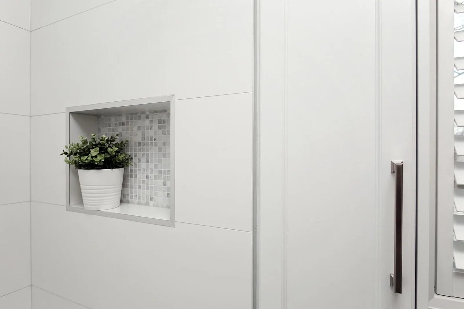 White shower surround with a recessed mosaic niche, clean glass enclosure, and a small potted plant.