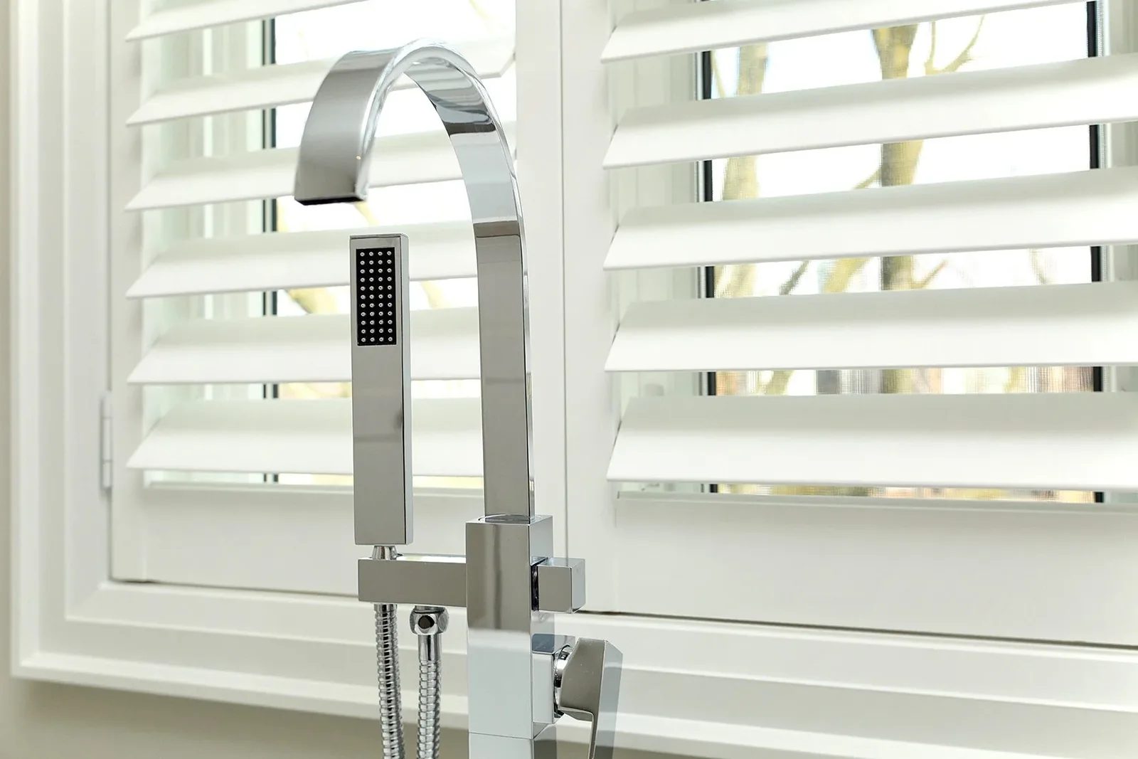 Polished chrome kitchen faucet and hand sprayer framed by white plantation shutters and bright window light.