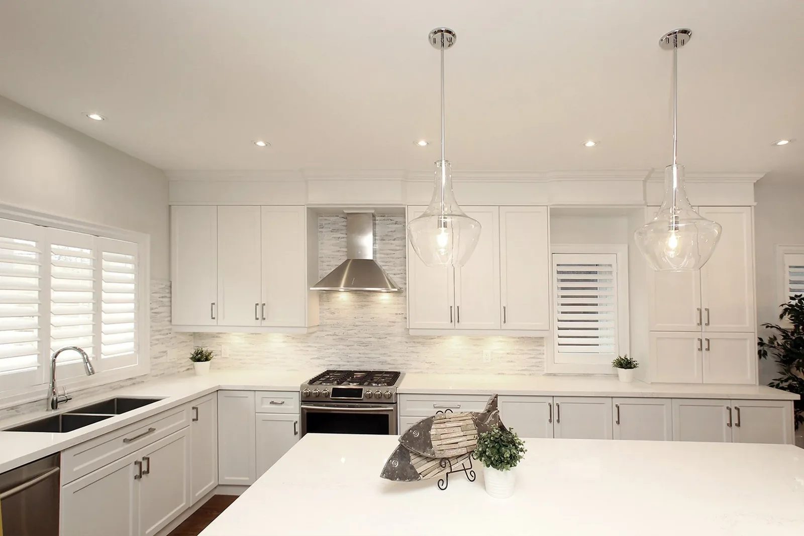Bright white kitchen with shaker cabinets, quartz island, glass pendants and plantation shutters.