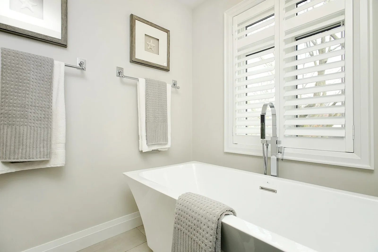 Bright bathroom with a freestanding tub, chrome filler, grey towels, and white shutters.