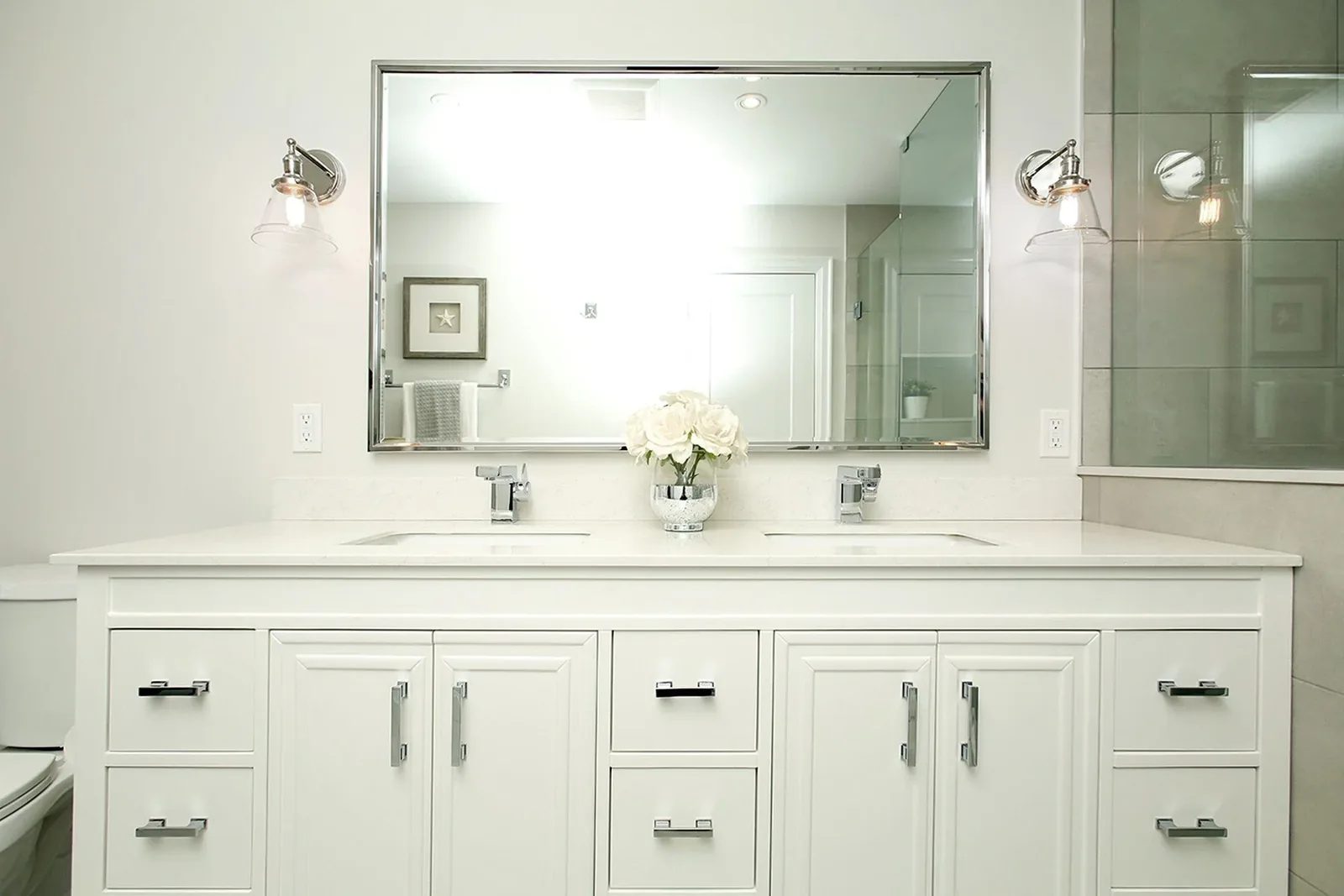 Bright white bathroom renovation with a double vanity, chrome sconces, a large mirror, and soft grey tile.
