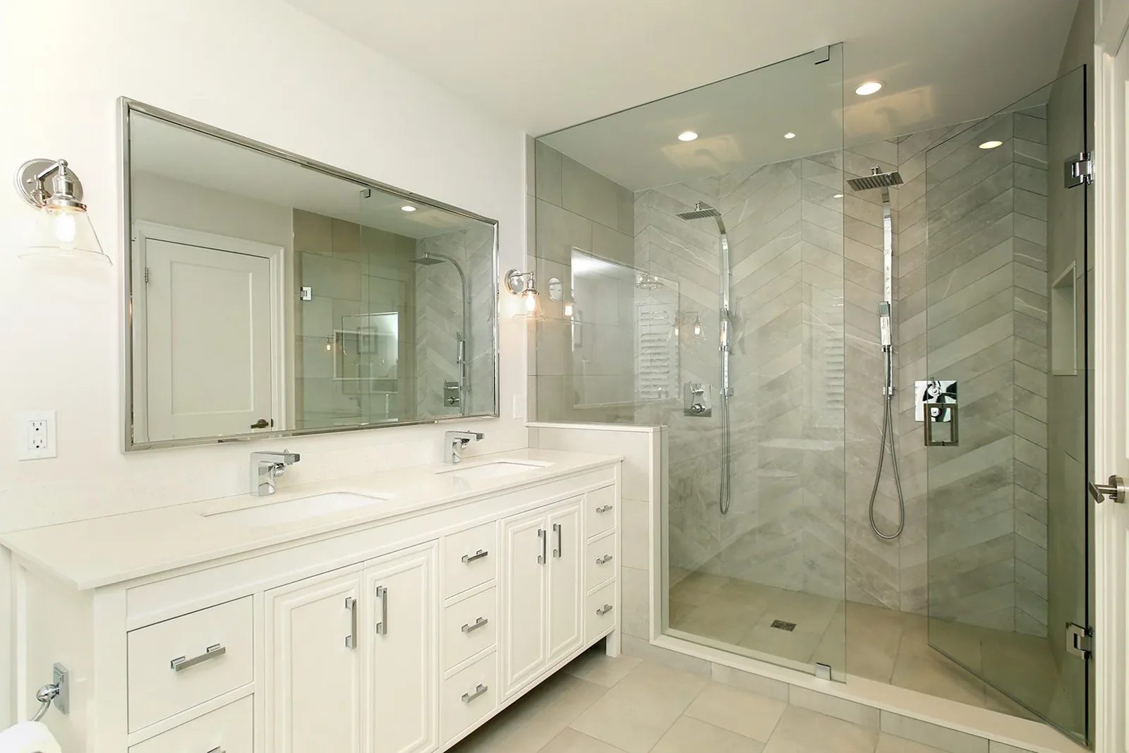 Bright white bathroom with a double vanity, framed mirror, and glass walk in shower with light grey tile.