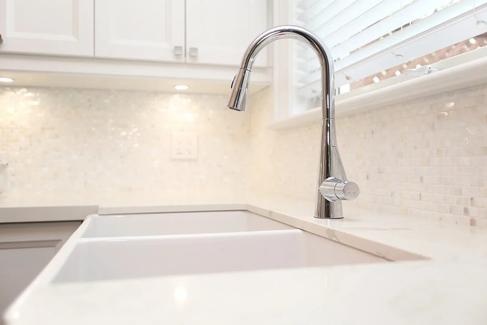 Polished chrome kitchen faucet over a white farmhouse sink with warm tile backsplash and bright window light.