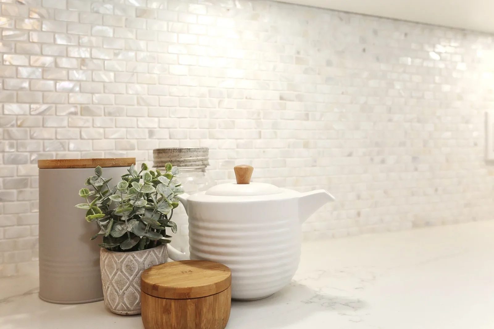Warm white kitchen backsplash detail with glossy tile, pale stone counter, and natural wood accents.
