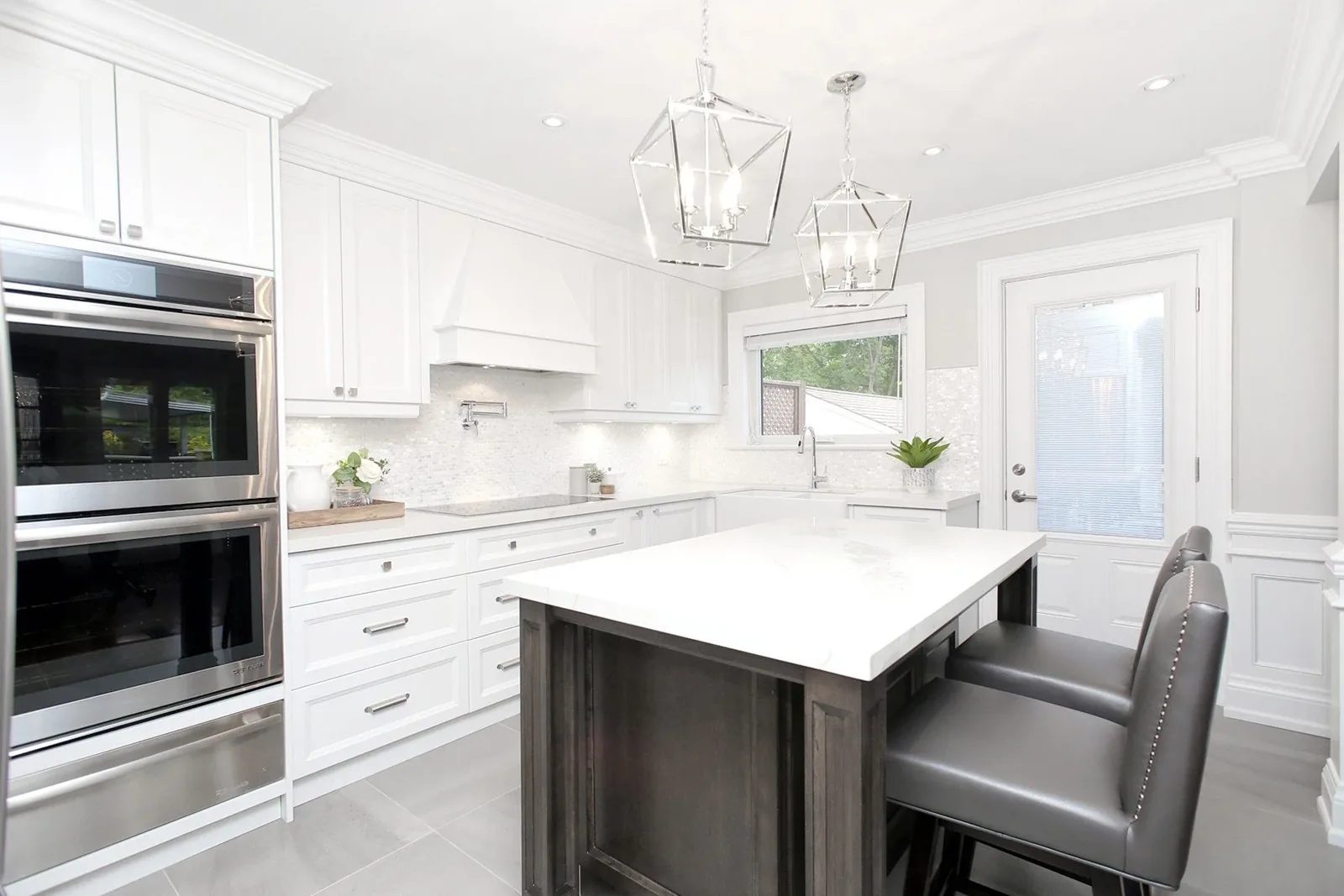 Bright white kitchen renovation with quartz island, wall ovens, grey stools and glass pendant lights.