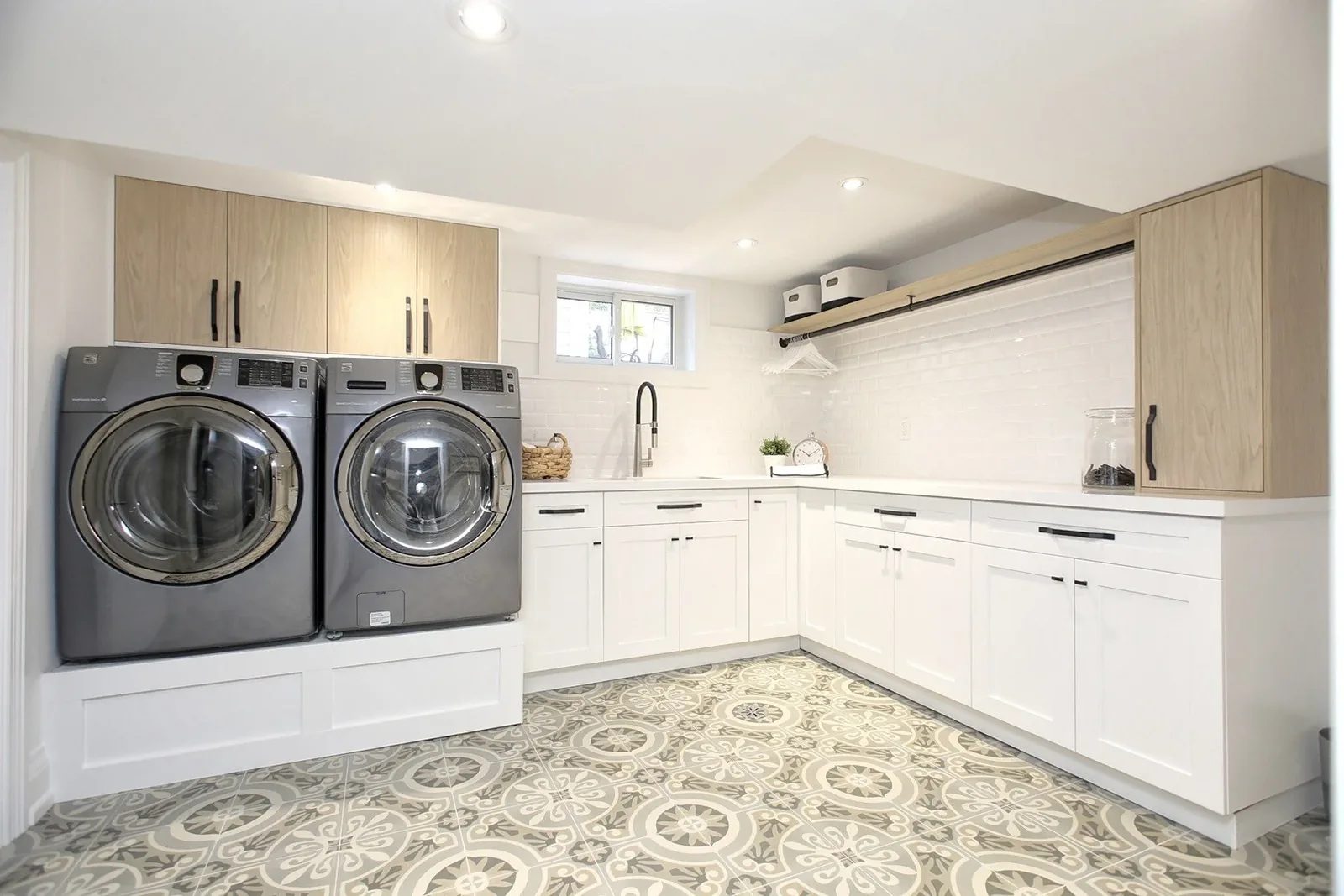 Bright basement laundry room with white shaker cabinetry, oak uppers, patterned tile and side by side machines.