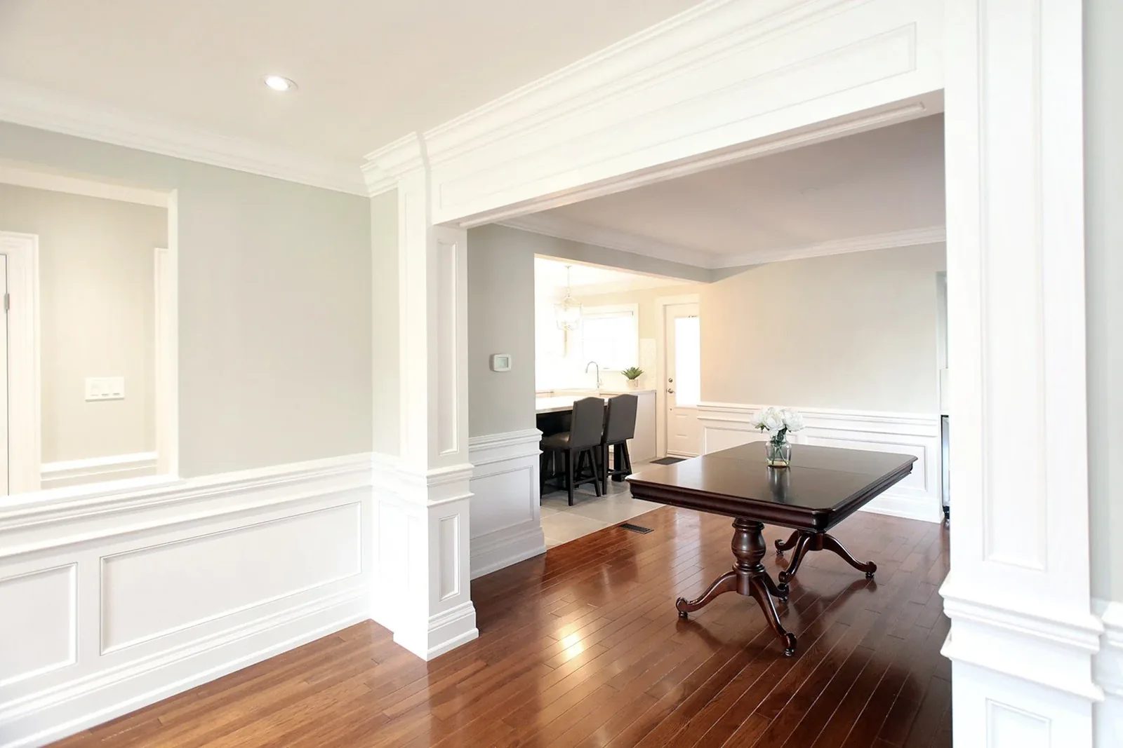 Bright dining room with white millwork, grey walls and dark hardwood, opening to a softly lit kitchen.