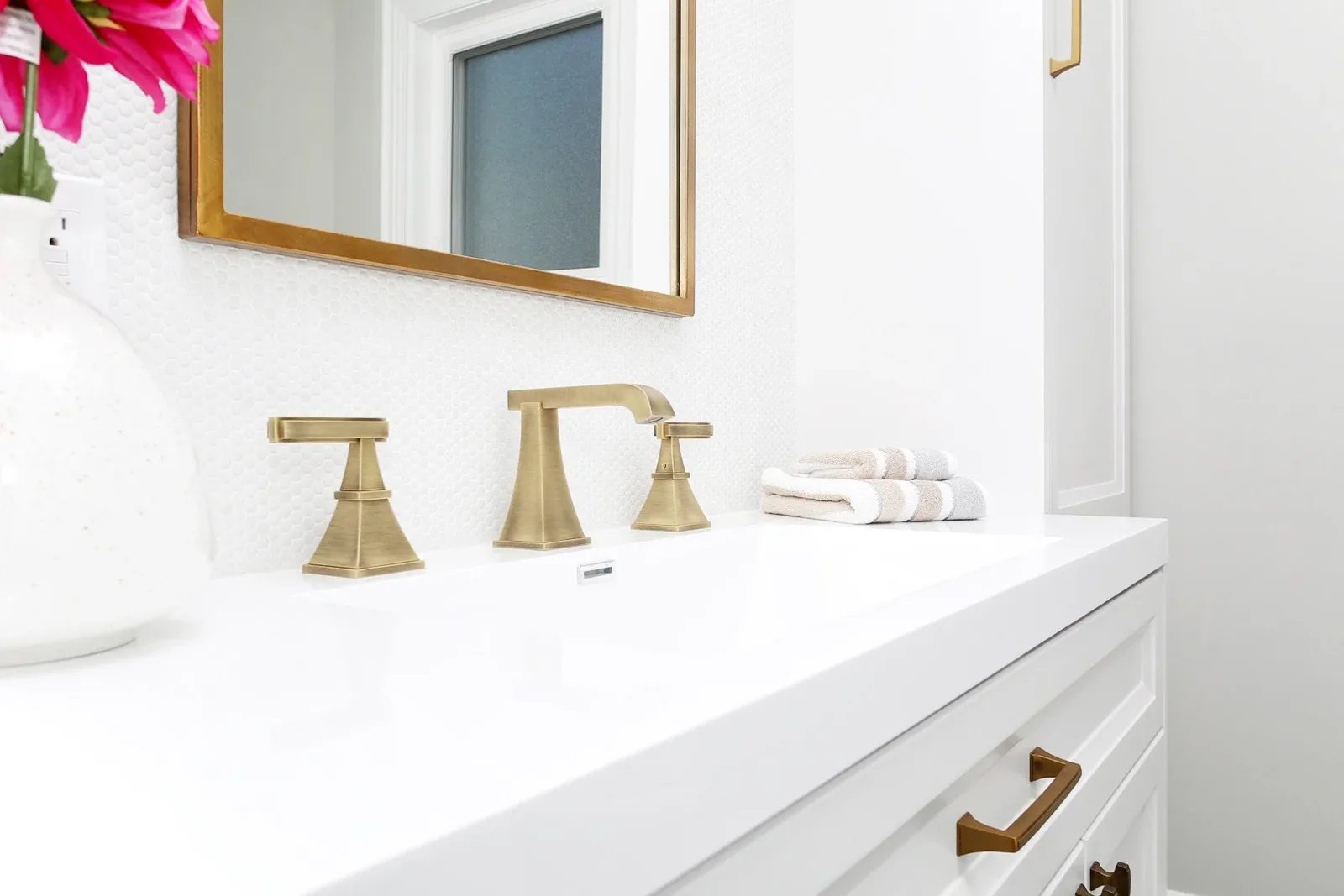 Bright bathroom vanity with warm white cabinetry, brass faucet, folded towels, and a gold framed mirror.