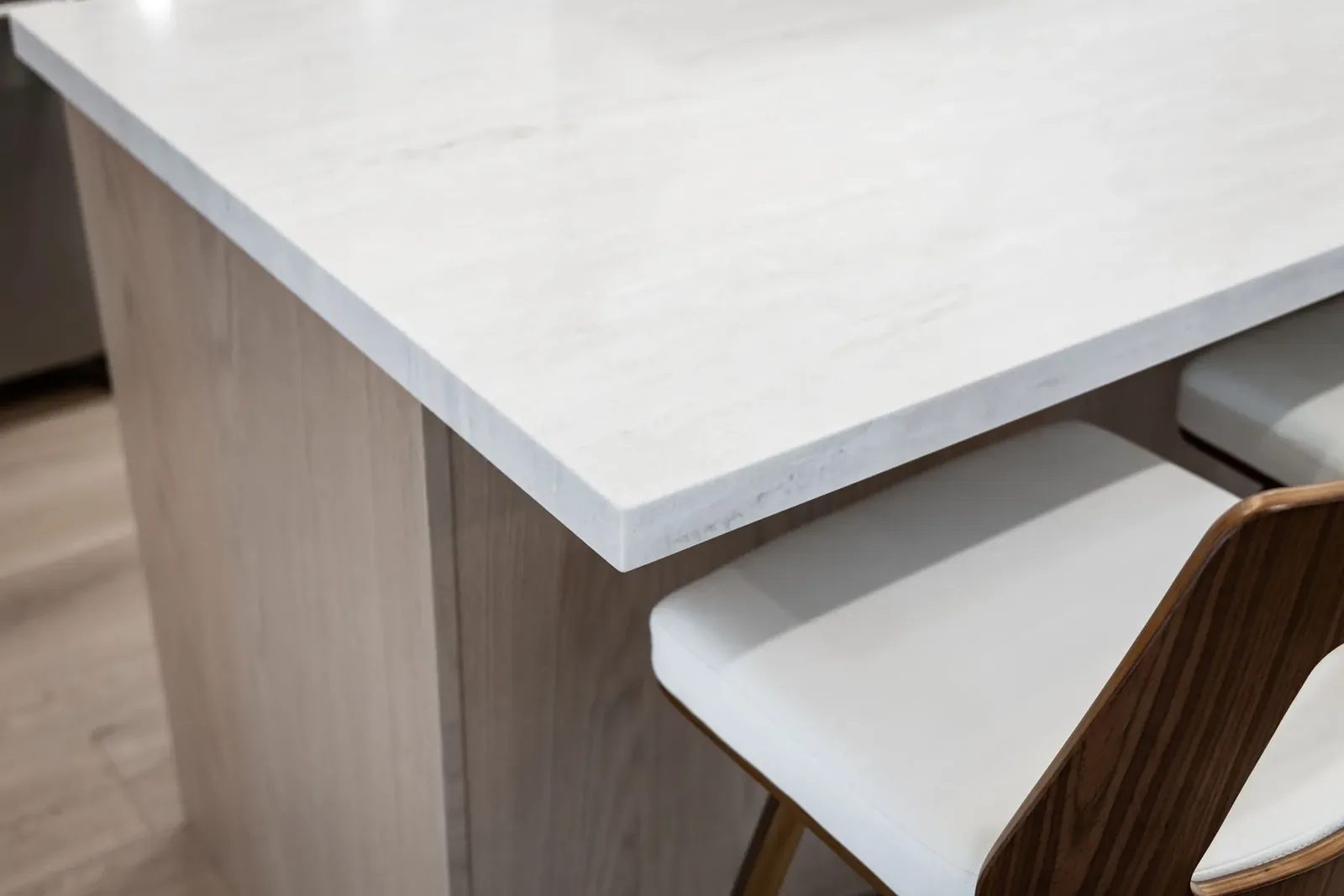 A white stone kitchen island with a pale wood base overhangs two stools with curved wood backs and white seats.