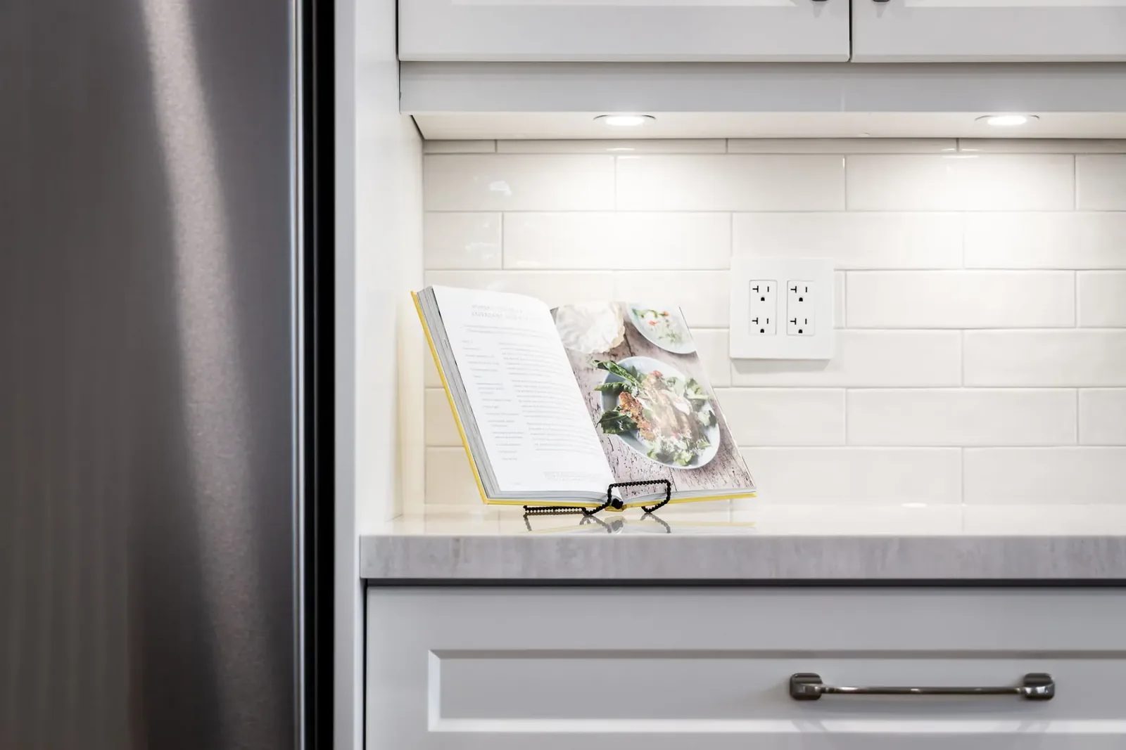 An open cookbook rests on a black stand beneath cabinet lights against a glossy white subway tile kitchen backsplash.