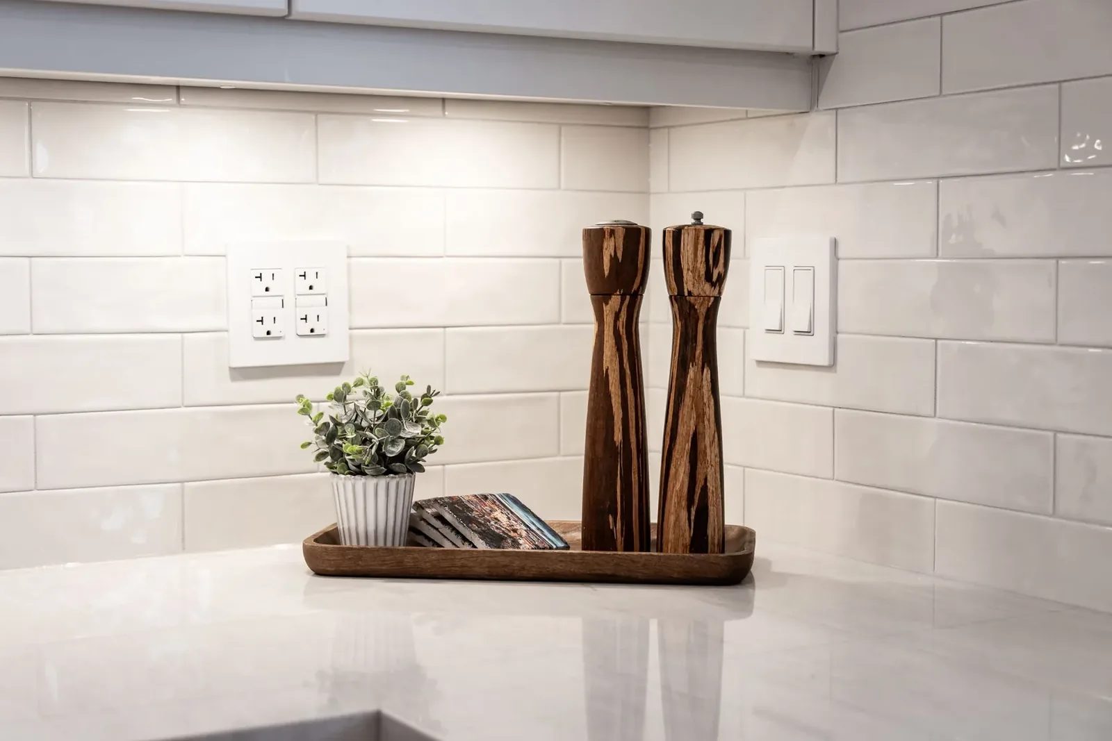 Two tall wood mills, a small potted plant and books sit on a tray against a glossy white subway tile kitchen backsplash.