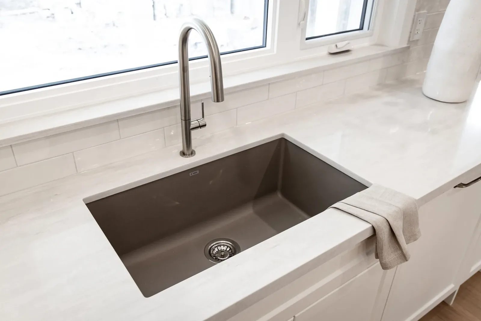 A deep grey undermount kitchen sink with a brushed metal gooseneck faucet sits below two windows in a white stone counter.