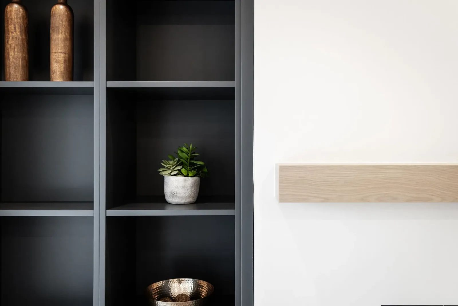 A small green plant in a grey pot and two bronze-coloured vessels sit on black built-in shelves beside the pale wood mantel in the family room.
