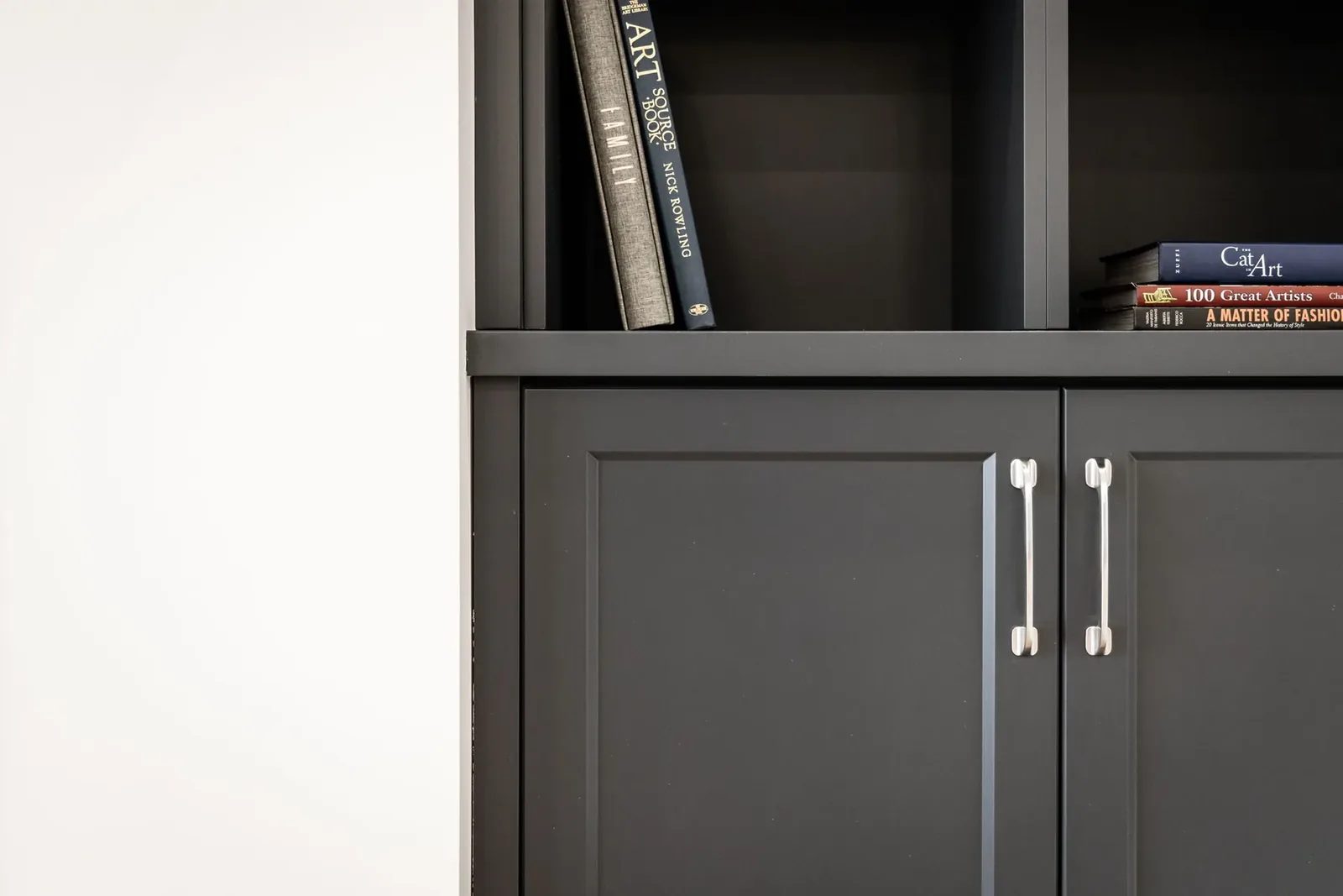 Black built-in shelving holds upright and stacked art books above matching cabinet doors with silver pulls in the family room.