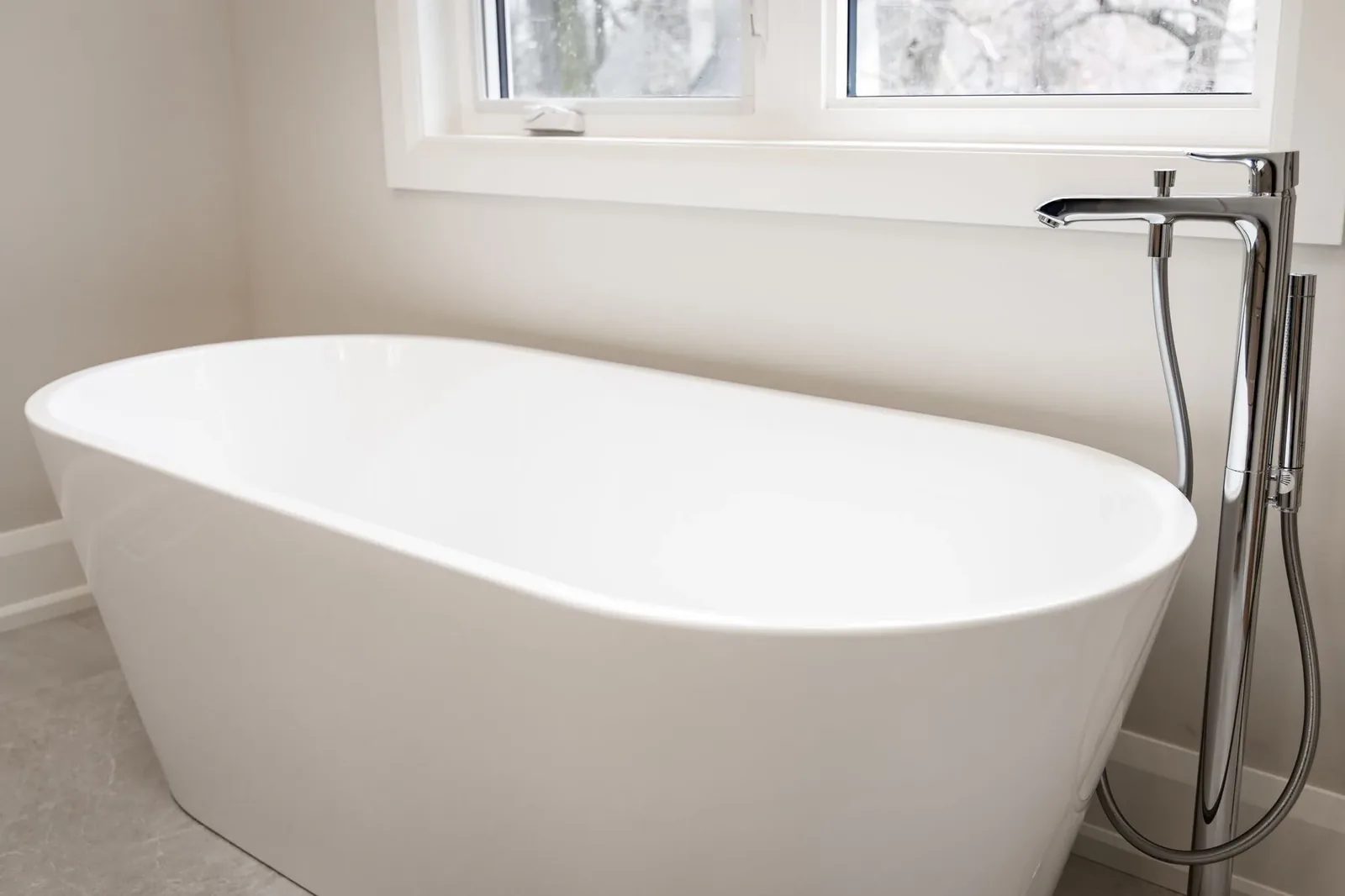 Freestanding oval tub with a chrome floor-mounted filler and hand shower beneath a bright window.