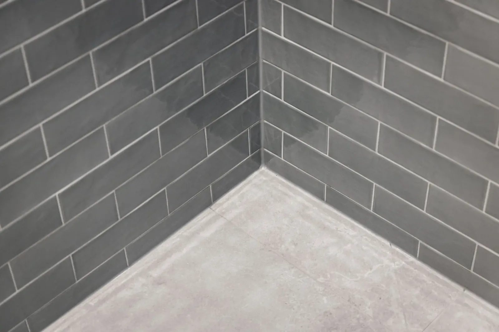 Glossy grey subway tiles meet large pale floor tiles at the lower corner of the hall bath shower.