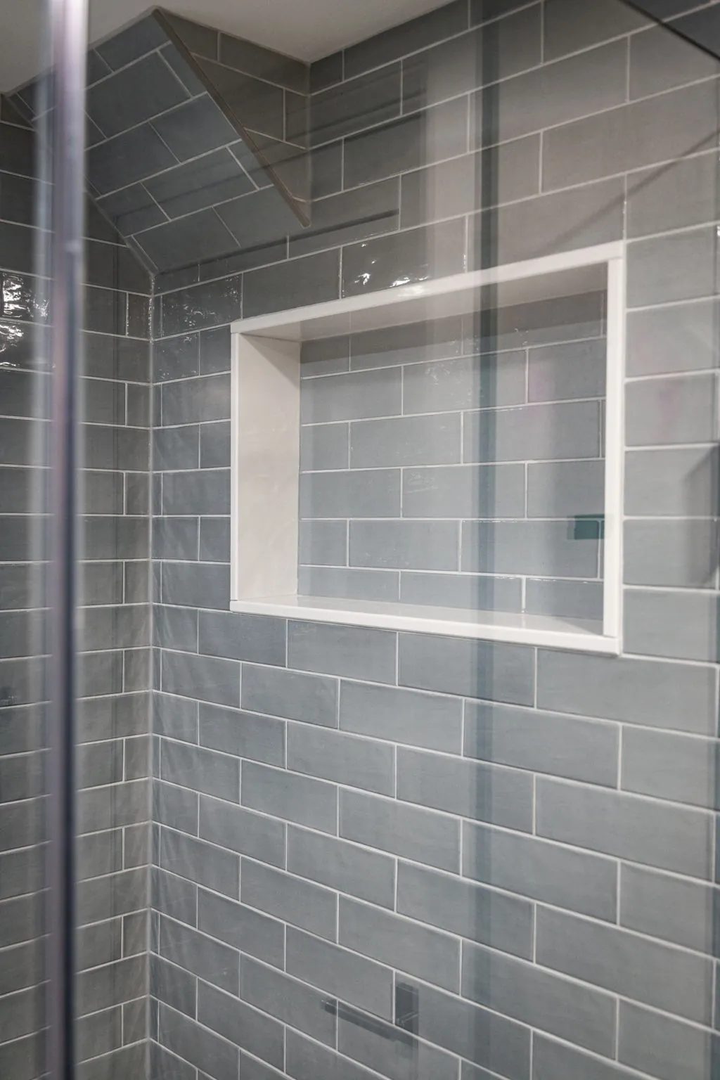A white rectangular shower niche is set into glossy grey subway tile behind a clear glass panel in the hall bath.