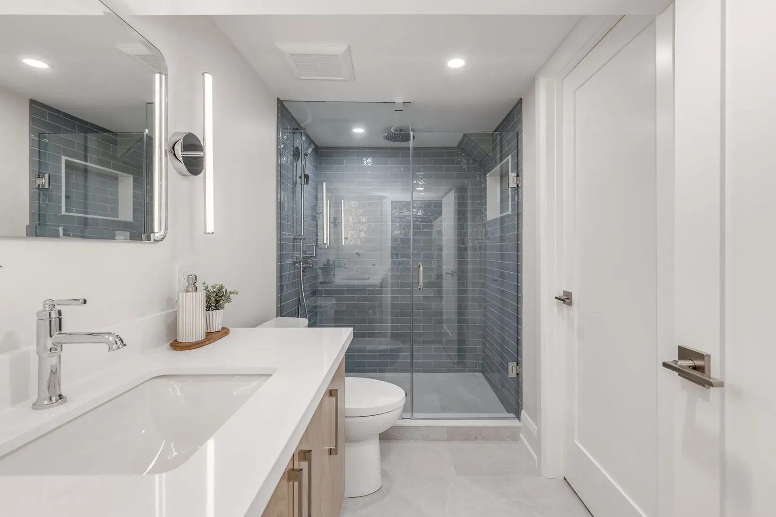 Second bathroom down its length, with the white counter and chrome faucet in the foreground and the blue-grey tiled shower behind glass beyond the toilet.