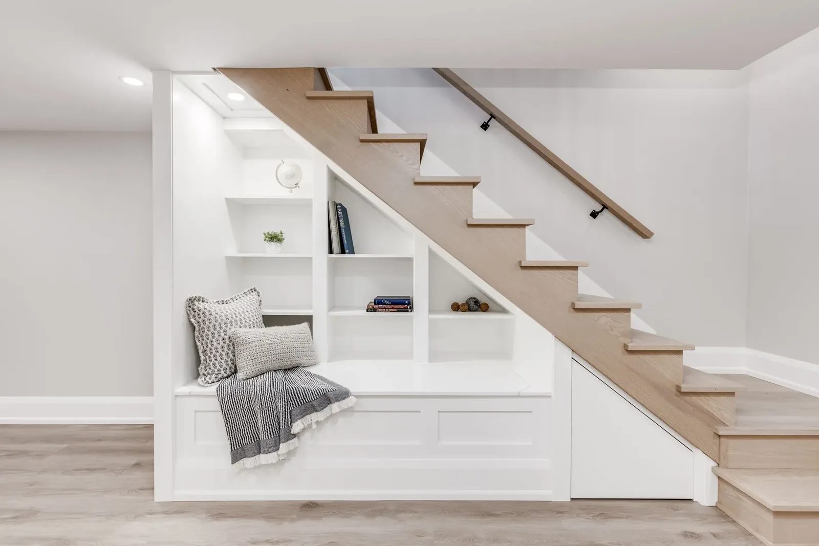 Under-stair reading nook with white built-in shelving, a cushioned bench and the oak treads climbing overhead.