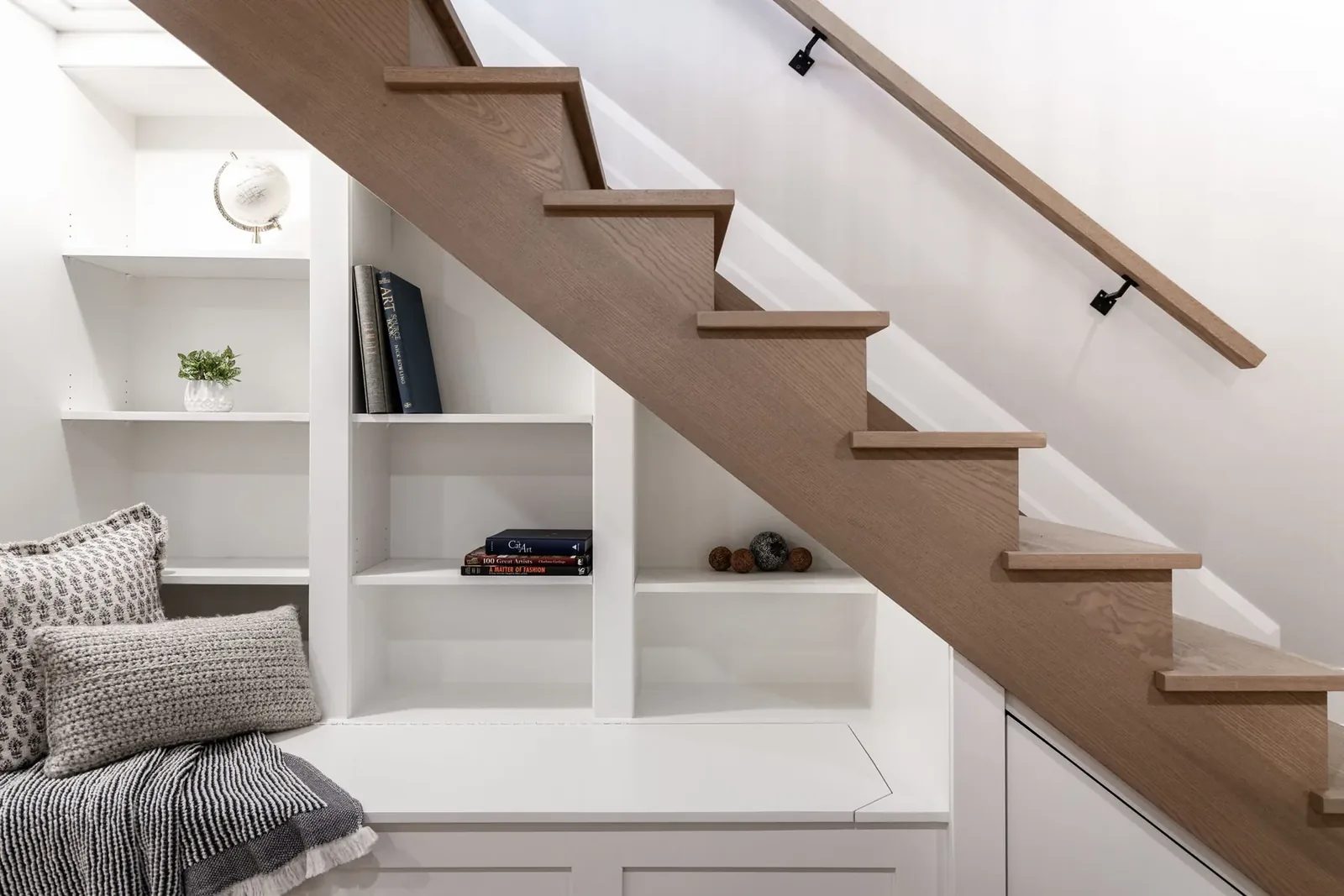 Reading nook up close, the white shelf bays and bench cushions tucked under the oak stair stringer.