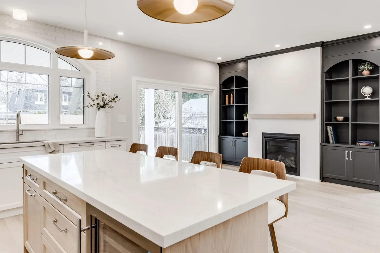 Across the quartz-topped island to the living side, the charcoal arched built-ins flanking the fireplace and the sliding glass door to the deck between them.