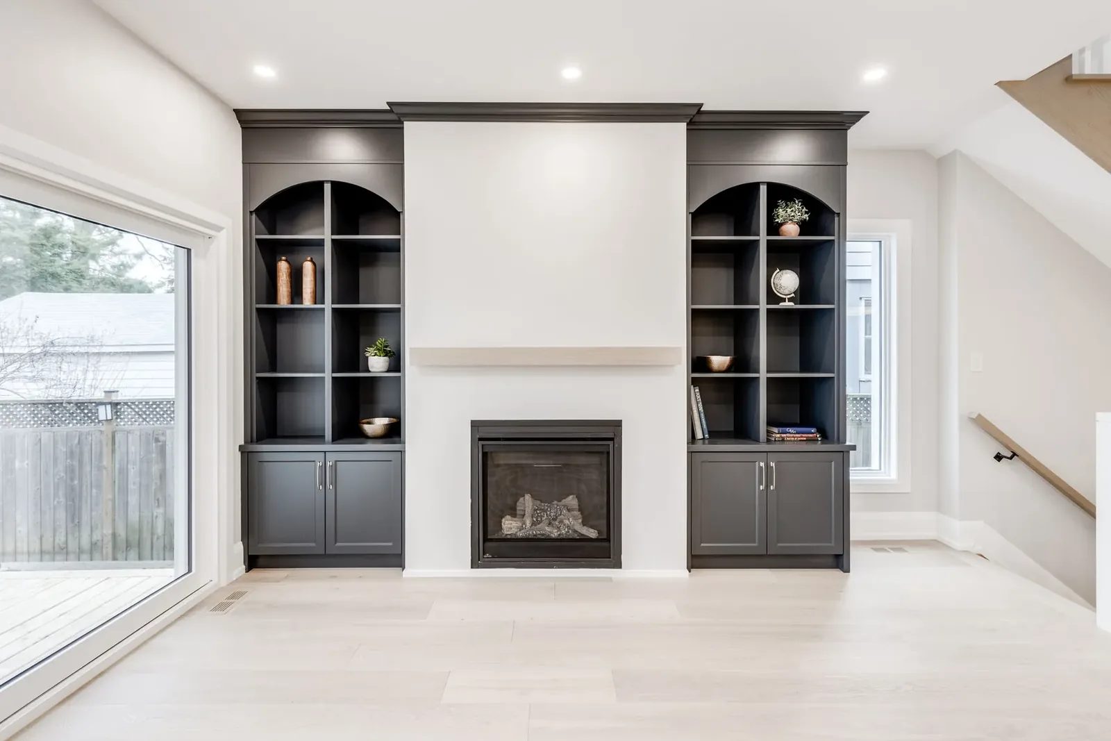 Charcoal built-ins straight on, arched upper bays over cabinet bases flanking the fireplace under a pale oak mantel shelf, with the stair beyond.