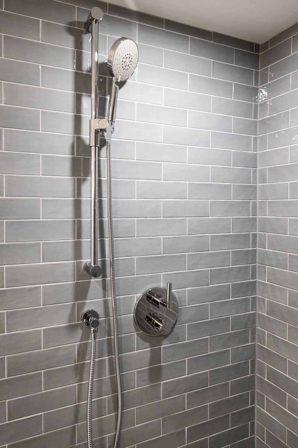 Shower corner of the second bathroom, a chrome hand shower on its riser bar beside a round thermostatic valve on blue-grey subway tile.