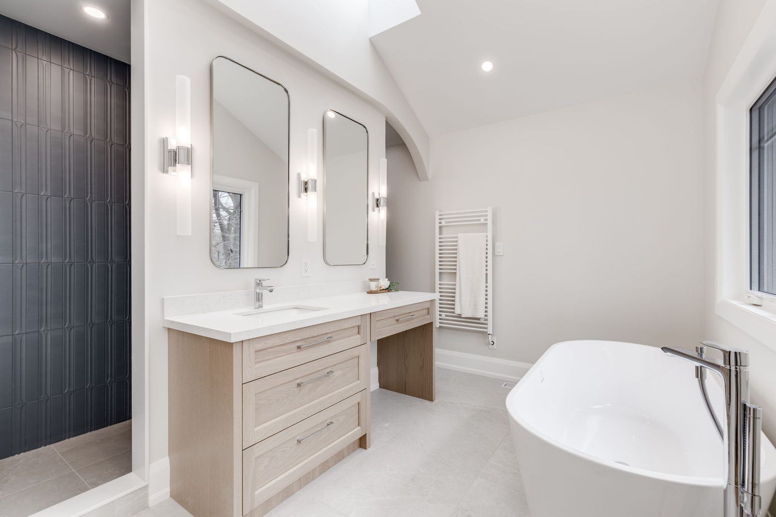 Primary ensuite with a light oak double vanity, twin mirrors and sconces, a dark fluted tile wet area and a freestanding tub under the skylight.