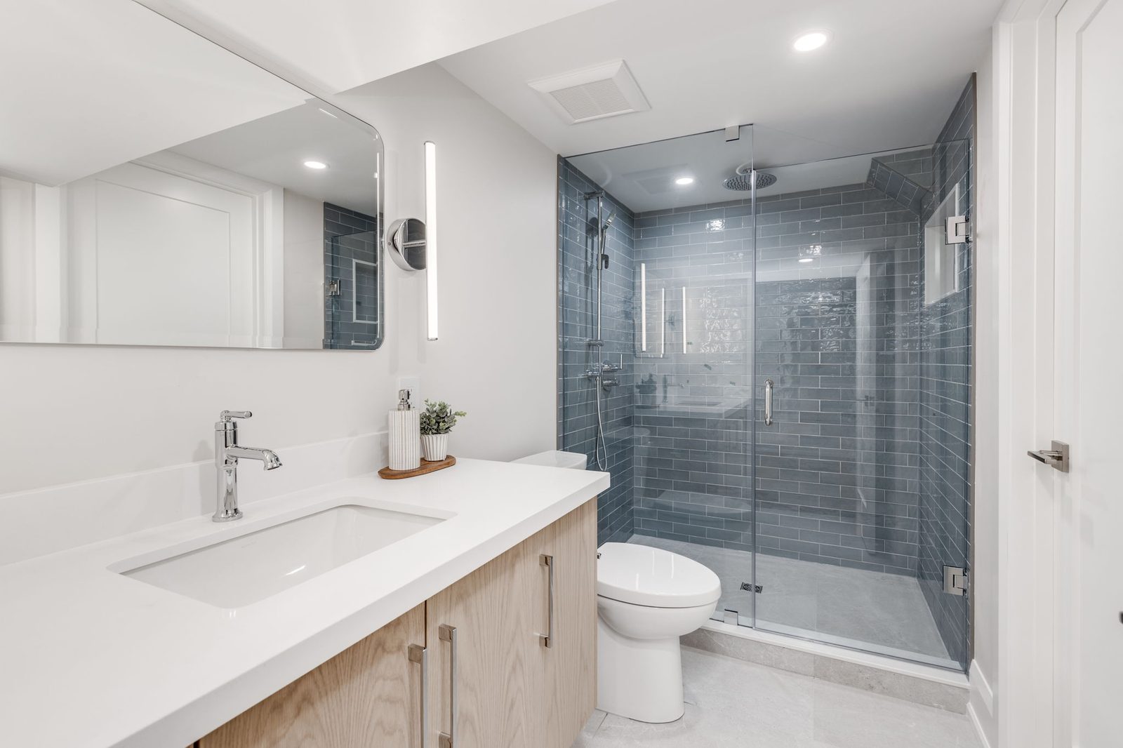 Light oak vanity with a white quartz counter and a glass-doored walk-in shower in blue-grey tile.