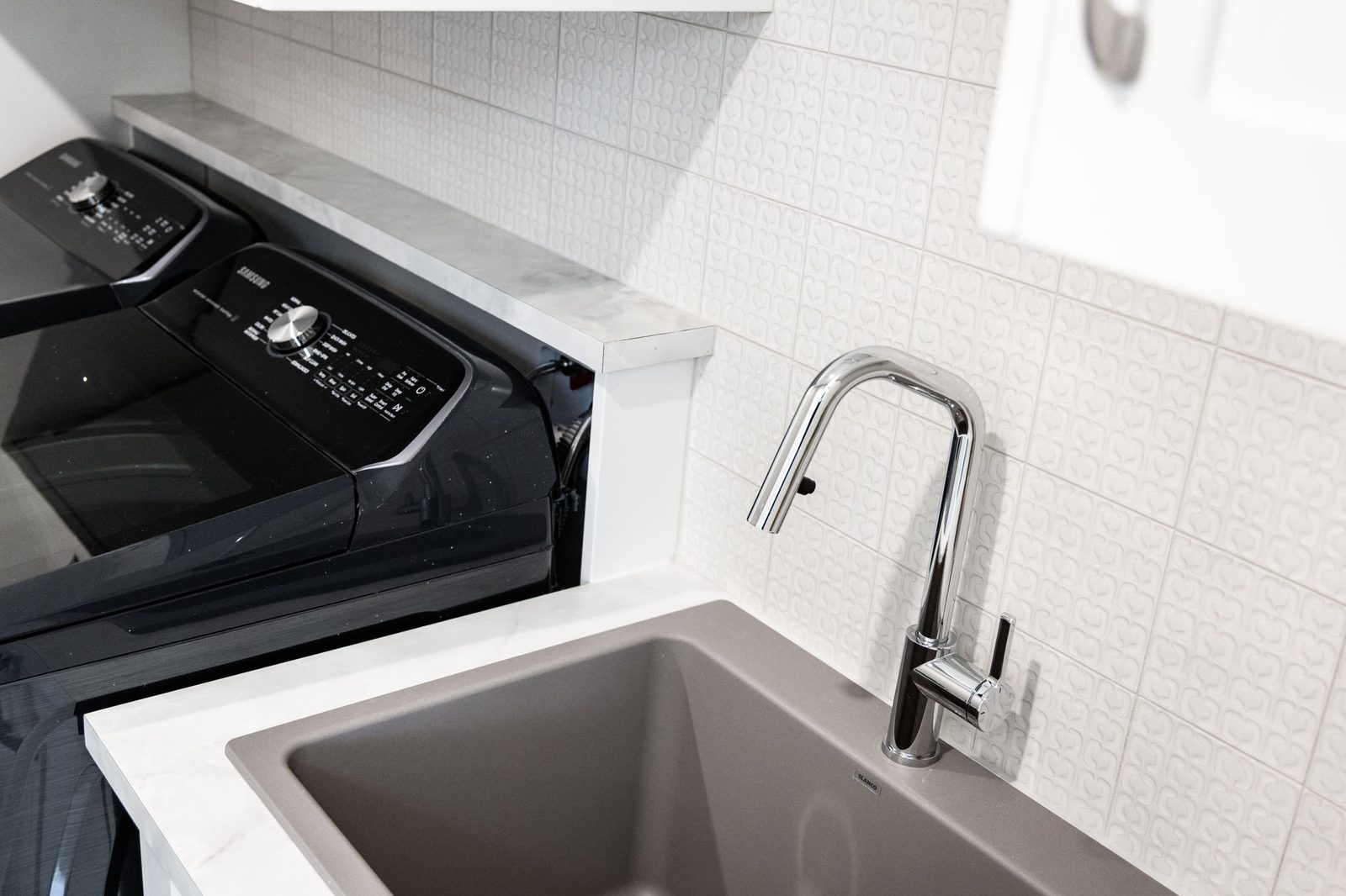 Laundry sink set into the counter beside the machines, against the textured white tile field.