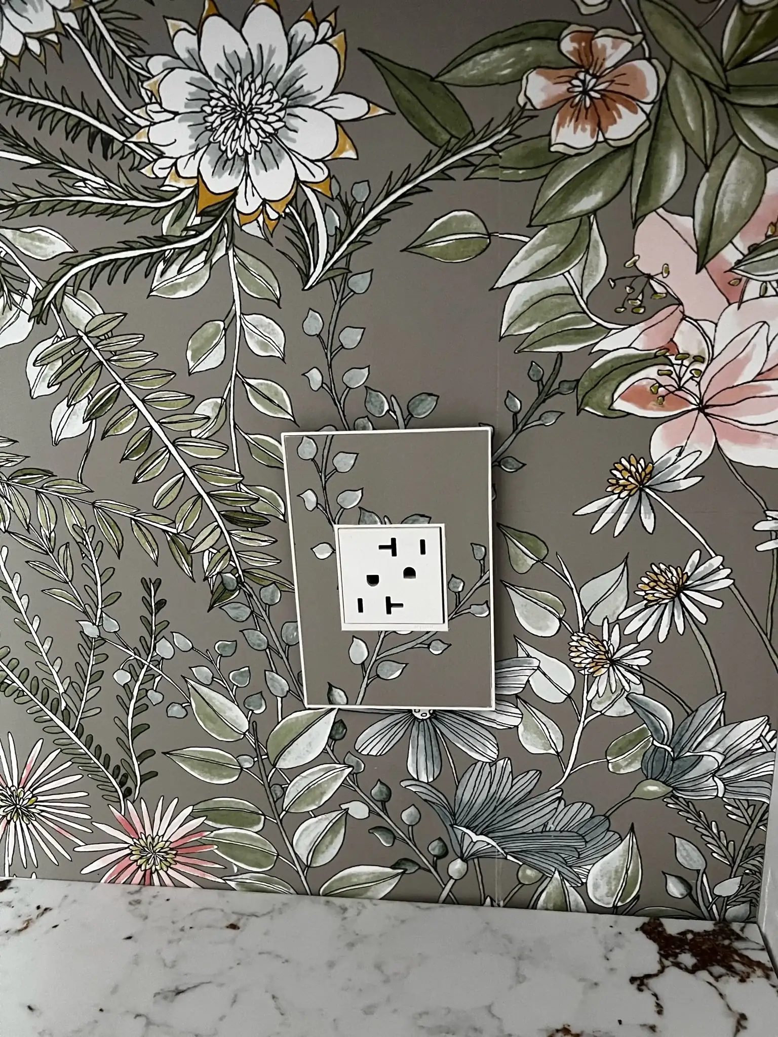 Botanical wallpaper surrounds a GFCI outlet above a white stone counter in a finished bathroom detail.