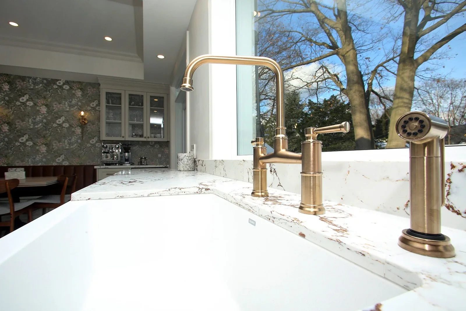 Brushed brass kitchen faucet over a white apron sink and veined stone counter beneath a large window.